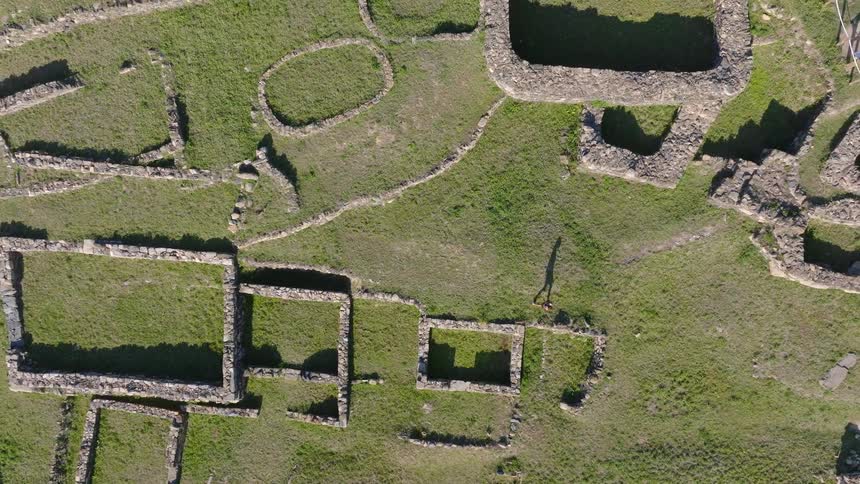 A slow top-down aerial pull-out reveals a person walking among the ancient stone foundations of a historic settlement on a sunny day