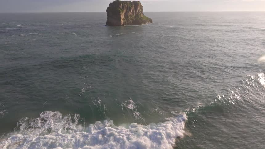 A powerful ocean wave crests in slow motion, pulling back to reveal a solitary sea stack at sunrise