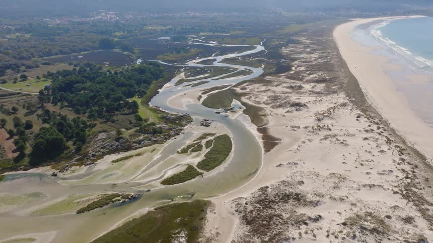 A stunning aerial view reveals a winding river estuary flowing through sandy dunes to meet the vast ocean along a picturesque coastline