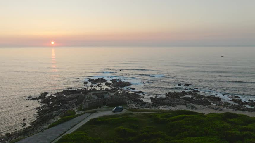 Aerial pan of a scenic rocky coastline featuring ancient ruins as the sun sets beautifully over the calm ocean