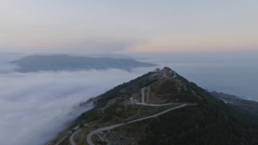 An aerial view reveals a mountain peak on the coast, with winding roads and communication towers rising above a sea of clouds at dawn