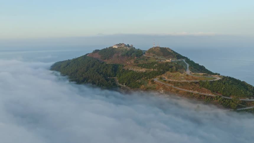 An aerial view captures a majestic mountain peak with winding roads rising above a sea of clouds at sunrise