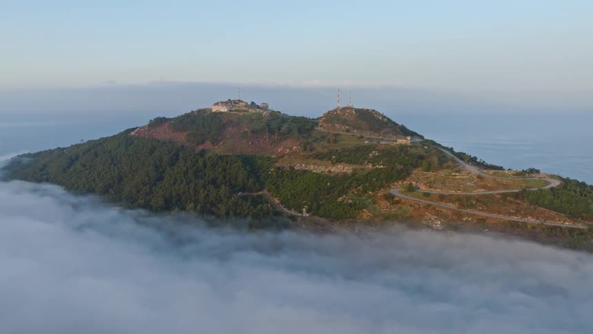 An aerial pullback reveals a scenic coastal mountain peak with a fortress and towers rising above a dense layer of morning fog