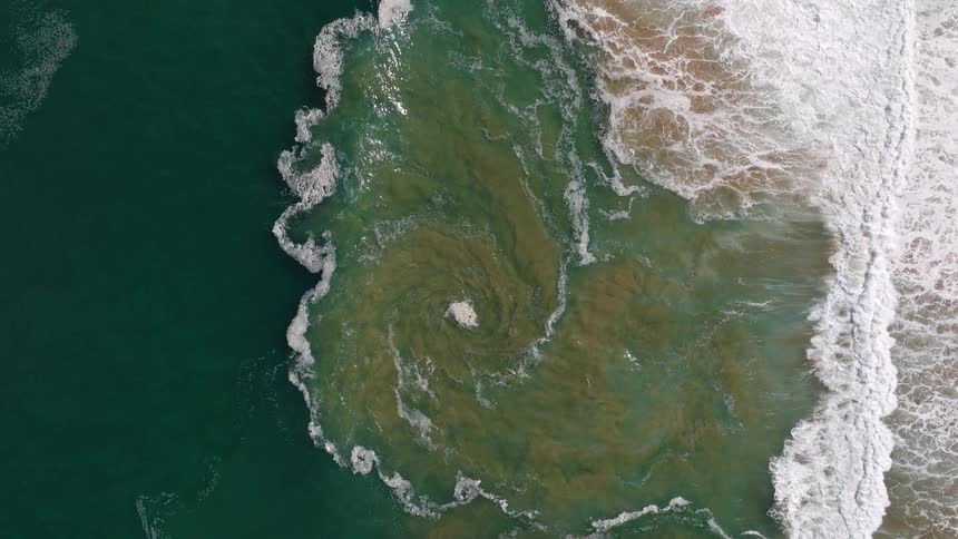 Top-down aerial view of a hypnotic ocean vortex created by crashing waves in slow motion