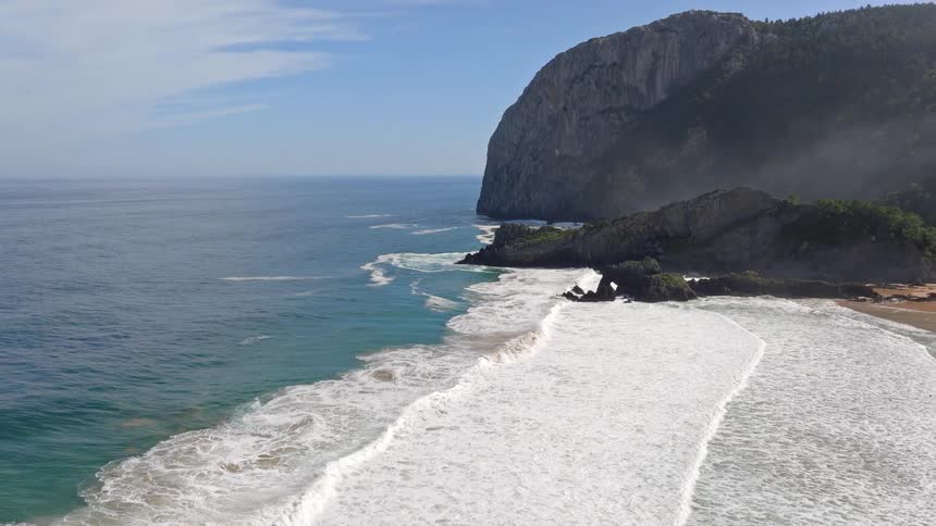 A serene, slow-motion aerial view of a secluded beach with waves gently rolling ashore next to a verdant, misty cliff