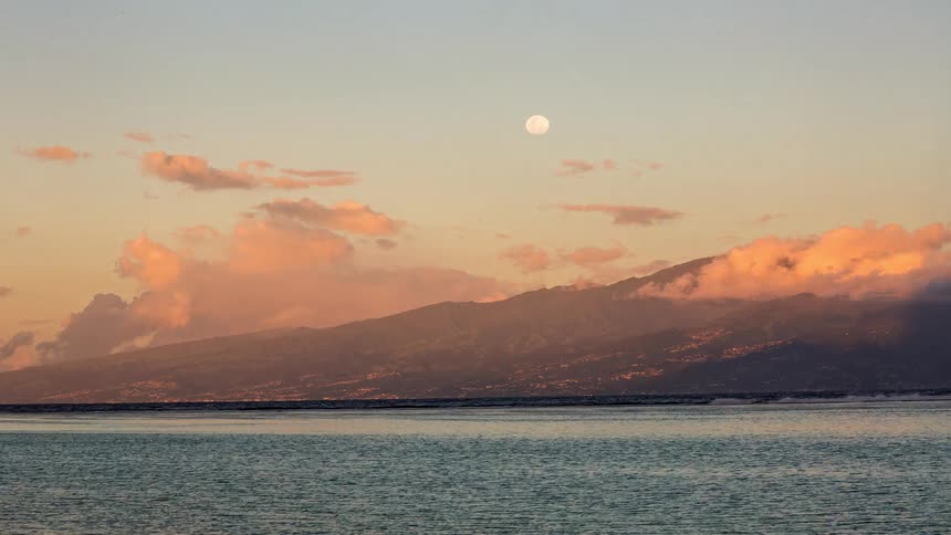 A picturesque full moon shines in the pastel-colored sky over a mountainous island and the tranquil sea at dusk