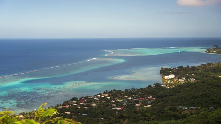 High angle time-lapse of a stunning tropical coastline with a village nestled in the jungle, overlooking a turquoise lagoon and coral reef