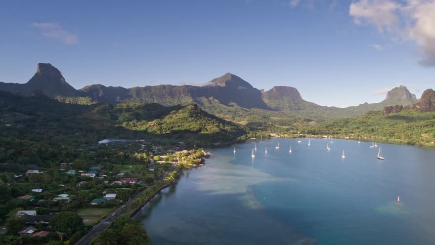Stunning aerial pull-back shot of sailboats in a tranquil turquoise bay at the base of lush volcanic mountains on a tropical island