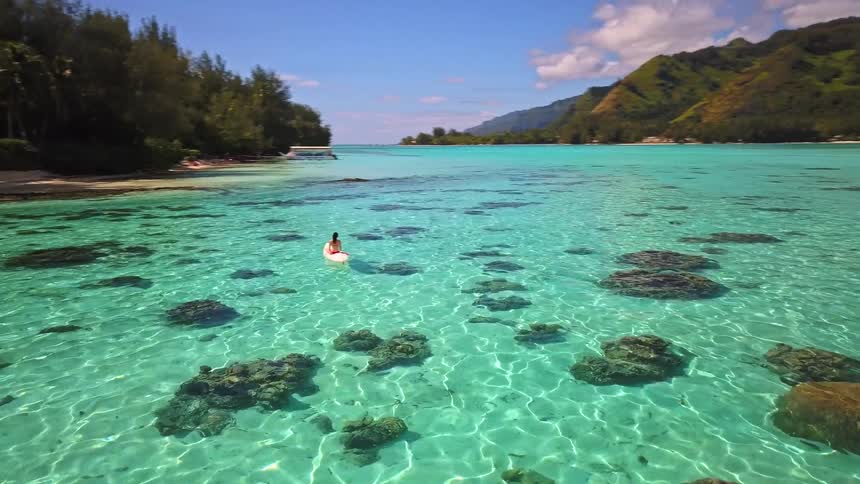 An aerial view captures a woman peacefully sitting on a paddleboard in a stunningly clear turquoise lagoon by a tropical island