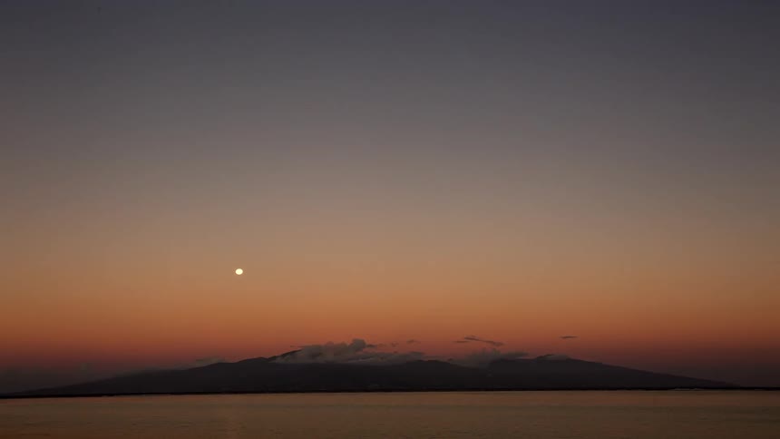 A serene timelapse captures the sunset over a distant island and calm ocean as the moon rises in the colorful sky