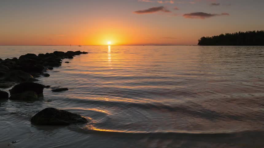 A serene time-lapse captures the sun setting over a calm tropical sea with gentle waves lapping on the sandy shore