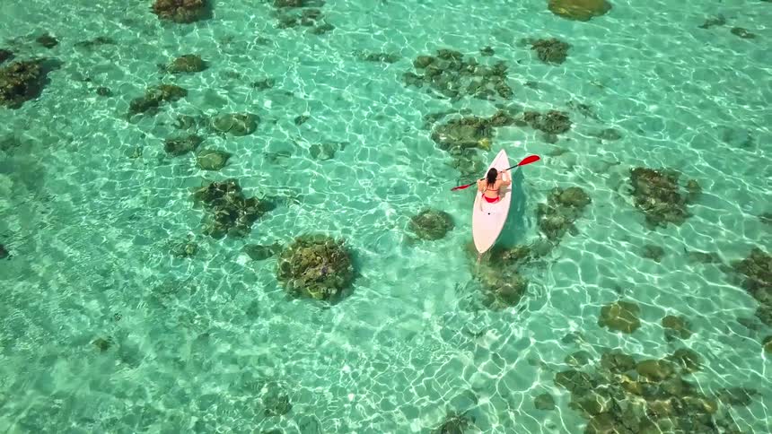 Stunning aerial view of a woman kayaking in a crystal clear turquoise lagoon, slowly revealing a lush tropical island