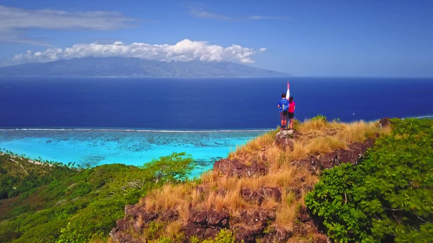 Aerial view rises over a couple on a hilltop with a flag to reveal a spectacular tropical island, turquoise lagoon, and luxury overwater resort