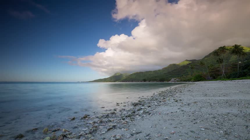 Serene long exposure timelapse captures a tropical beach with calm turquoise waters and clouds moving over lush green mountains