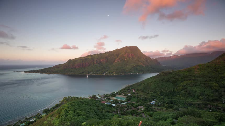 An epic day-to-night timelapse shows golden hour light fading to dusk over a lush tropical island bay with sailboats and a rising moon