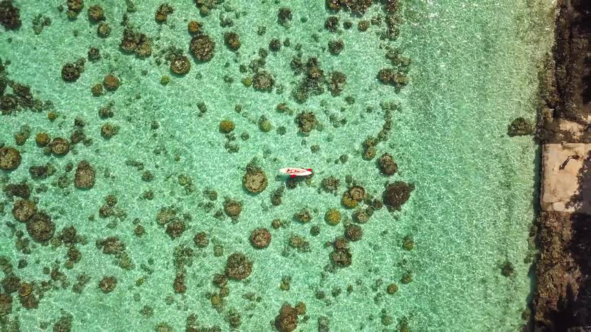 A stunning aerial view pulls back to reveal a woman in a bikini relaxing on a surfboard, floating peacefully over a vibrant coral reef in a crystal-clear turquoise lagoon