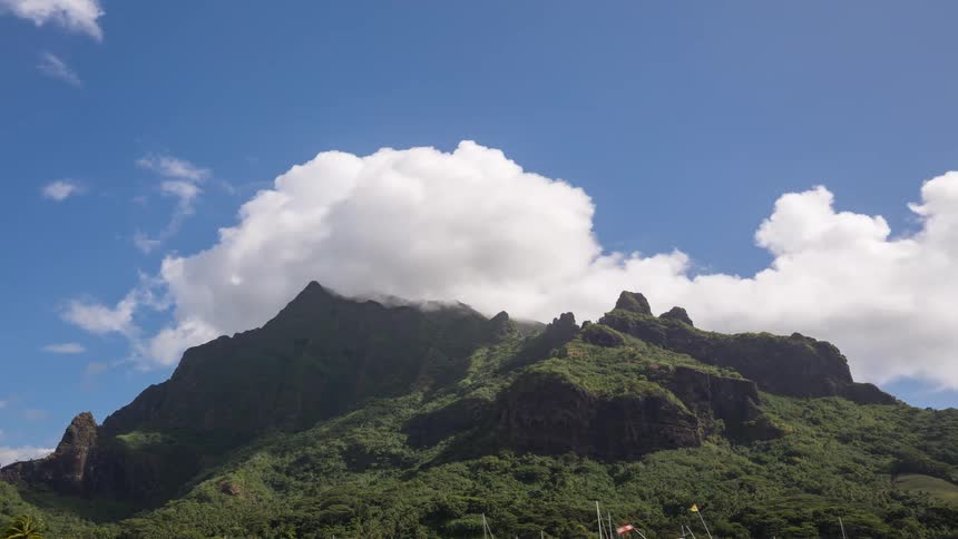 A timelapse of fluffy white clouds moving across a bright blue sky over a majestic, lush green tropical mountain