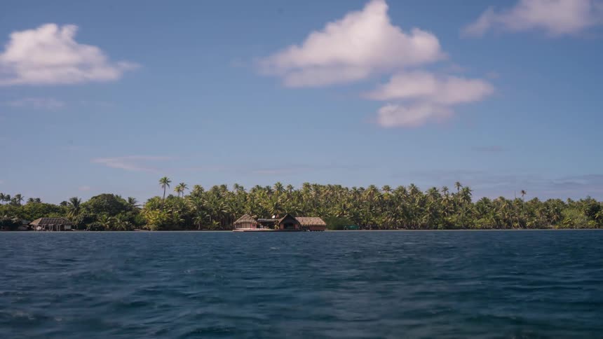 A tranquil view from the ocean of a remote tropical island covered in lush palm trees with traditional huts on the shoreline