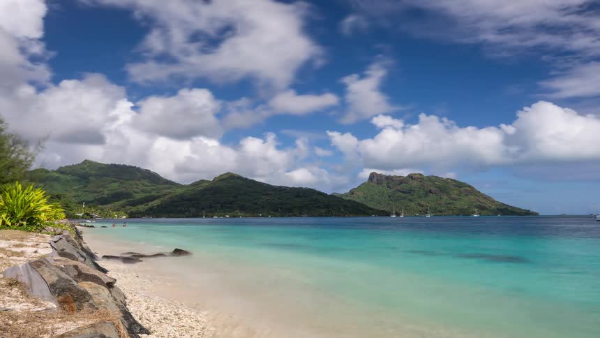 A stunning time-lapse of a pristine tropical beach with turquoise water, lush green mountains, and fast-moving clouds across a vibrant blue sky