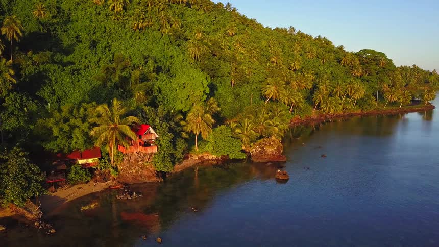 Drone flies away from a hidden oceanside bungalow nestled in a lush tropical jungle during a beautiful sunset
