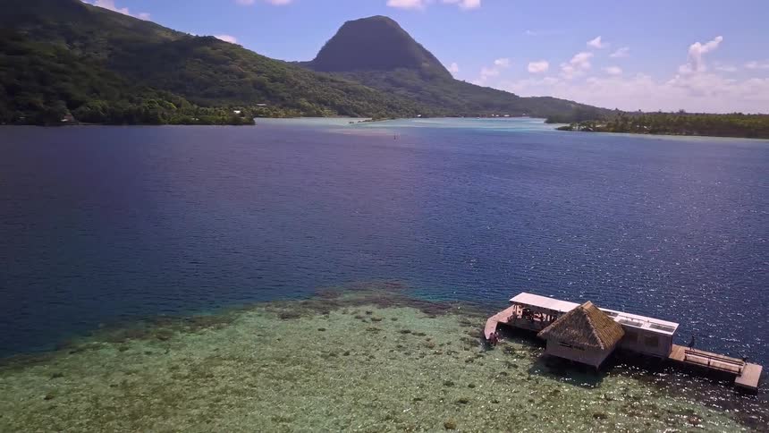 A slow aerial pullback reveals a beautiful overwater bungalow situated on a vibrant coral reef in a deep blue tropical lagoon