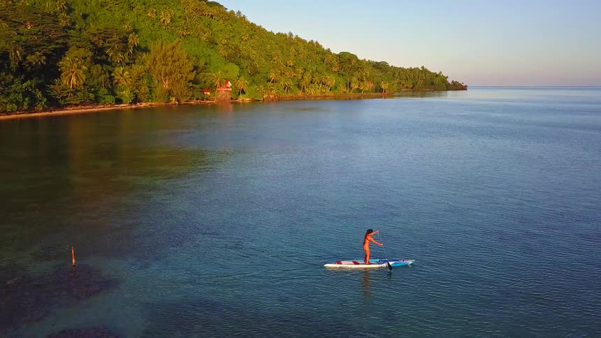 An aerial view shows a woman in a bikini stand-up paddleboarding on calm turquoise water along a lush tropical island coastline at sunrise
