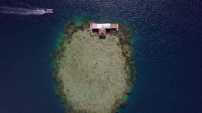 An aerial top-down shot slowly descends over a secluded coral reef with a single overwater bungalow in the middle of the deep blue ocean as a small boat passes by