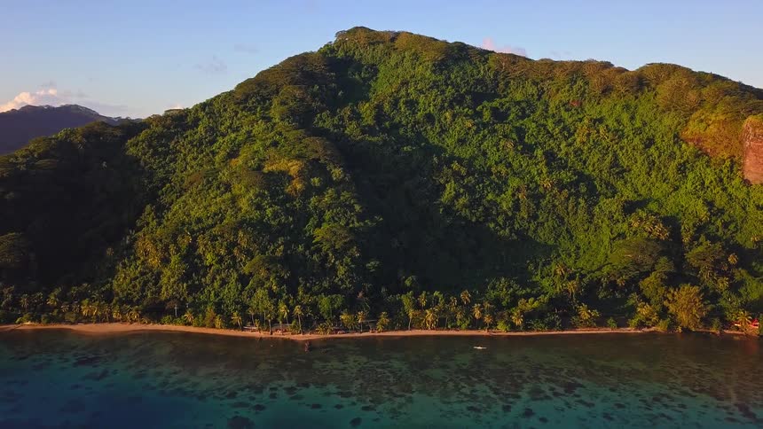 Aerial pullback reveals a lush, mountainous tropical island with a palm tree-lined beach and coral reef during a golden hour sunset