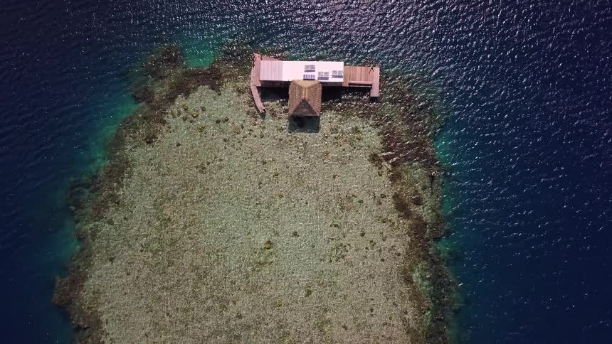 An aerial drone shot slowly pulls away from a secluded overwater bungalow situated on a beautiful coral reef in the middle of a turquoise ocean