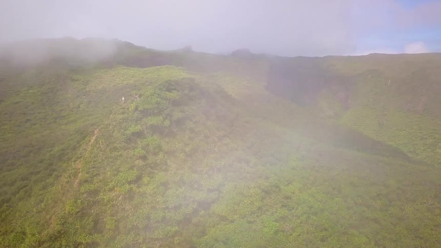 Aerial drone footage pushes through misty clouds over a lush green mountain ridge revealing hikers on an epic trail