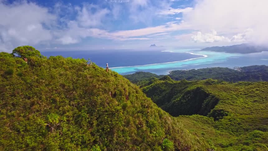 An epic aerial flight over a lush green mountain ridge reveals a hiker enjoying the breathtaking panoramic view of a tropical island lagoon