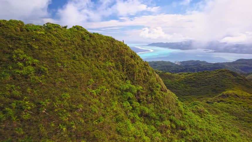 Inspiring aerial footage pulls back from hikers on a lush green mountain ridge, revealing a breathtaking view of a tropical island and its turquoise lagoon