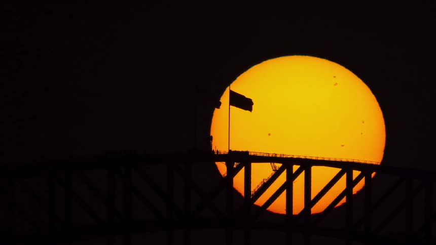A dramatic time-lapse shows the large orange sun setting behind the silhouette of the Sydney Harbour Bridge