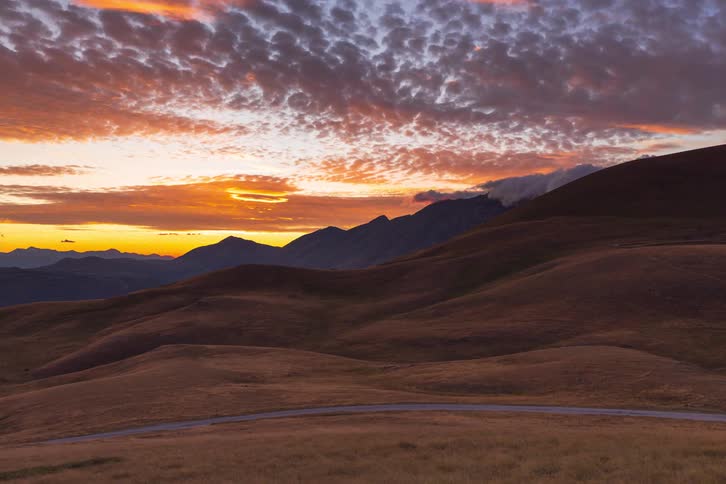 A dramatic timelapse shows a vibrant sunset over rolling mountain hills as a lone car drives along a scenic road into the fiery, colorful dusk