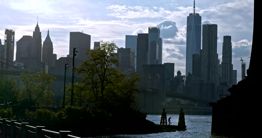 Silhouetted figures stand on a pier across the East River from the iconic downtown Manhattan skyline and Brooklyn Bridge