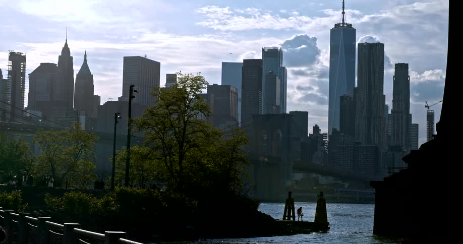 Silhouetted figures play on a pier along the East River with the iconic Lower Manhattan skyline and Brooklyn Bridge in the background