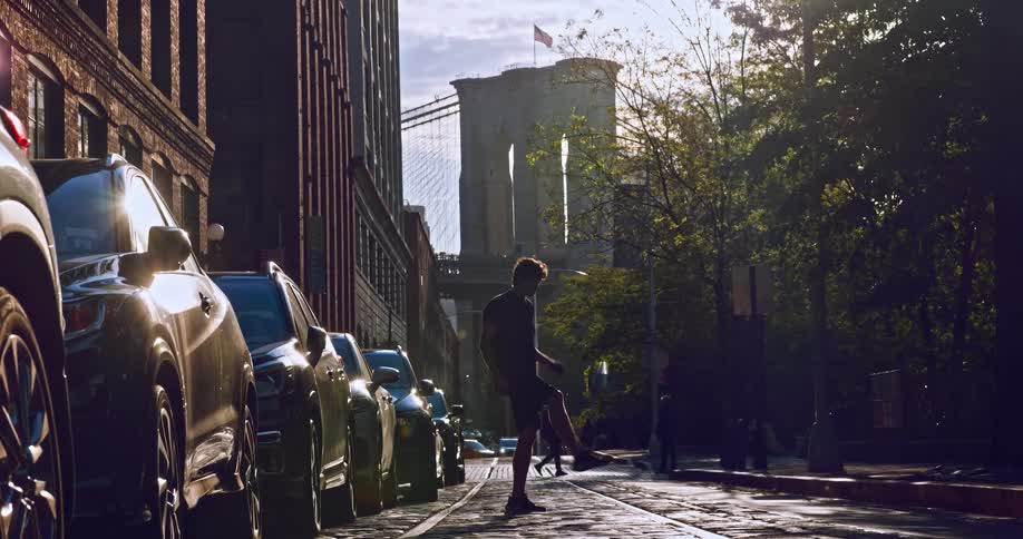 A young man skillfully juggles a soccer ball in slow motion on a cobblestone street in DUMBO, with the iconic Brooklyn Bridge in the background