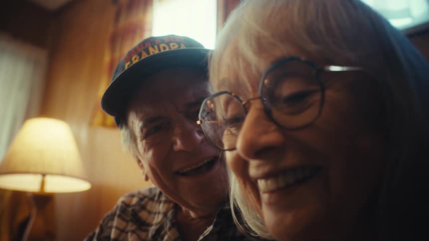 An intimate close-up shows a smiling elderly couple sharing a happy moment, with the man wearing a 'World's Best Grandpa' hat