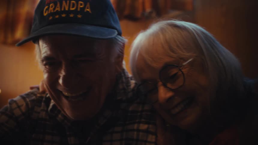 A heartwarming close-up of a happy elderly man in a 'World's Best Grandpa' hat as his wife lovingly rests her head on his shoulder