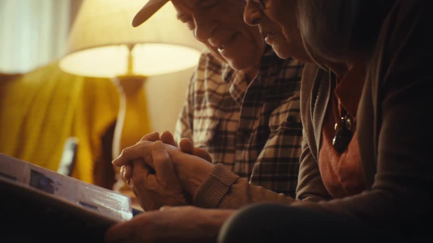 A loving senior couple holds hands and smiles warmly as they look through a photo album together in their cozy home