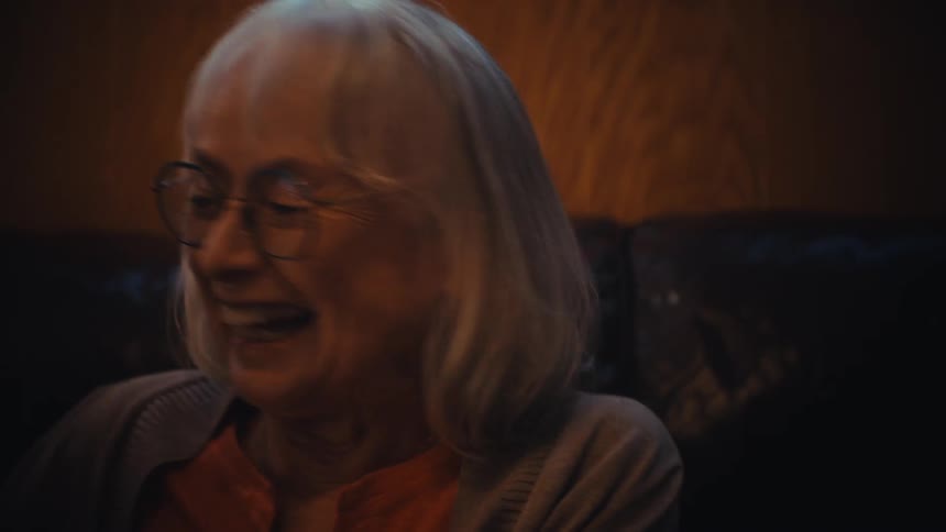 A loving elderly couple, with the man wearing a 'World's Best Grandpa' hat, laughs while reminiscing over old photos in an album