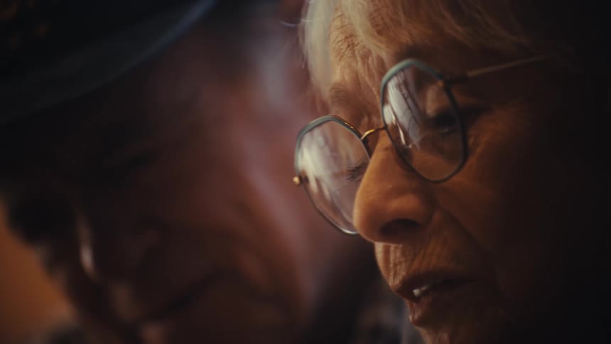 An intimate close-up of an elderly woman with glasses smiling and talking to her husband