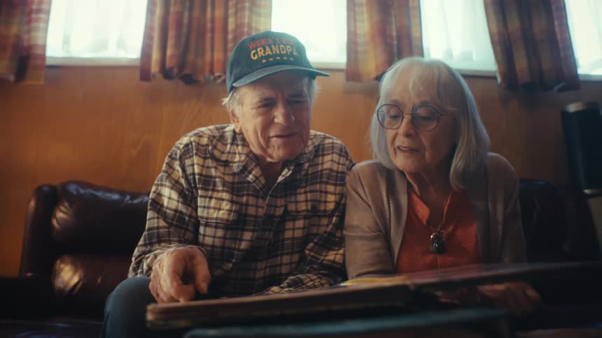 A senior couple, with the man wearing a 'World's Best Grandpa' hat, reminisces together while looking through a photo album