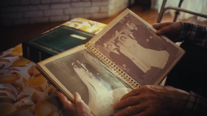 An elderly person reminisces while flipping through the pages of a vintage photo album, stopping on their old wedding pictures