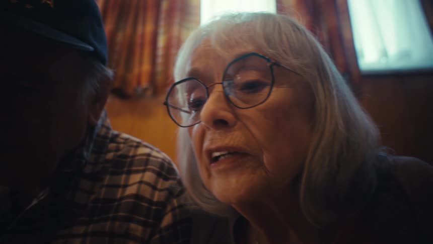An elderly couple, a grandpa in a 'World's Best Grandpa' hat and his wife, react with emotion and surprise while looking at a screen together