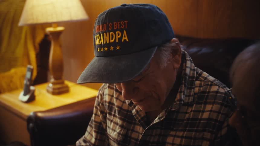 A close-up shot of an elderly wife comforting her emotional husband wearing a 'World's Best Grandpa' hat