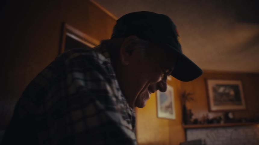 A smiling elderly grandpa in a plaid shirt and baseball cap uses a walker to slowly move through his warmly lit home