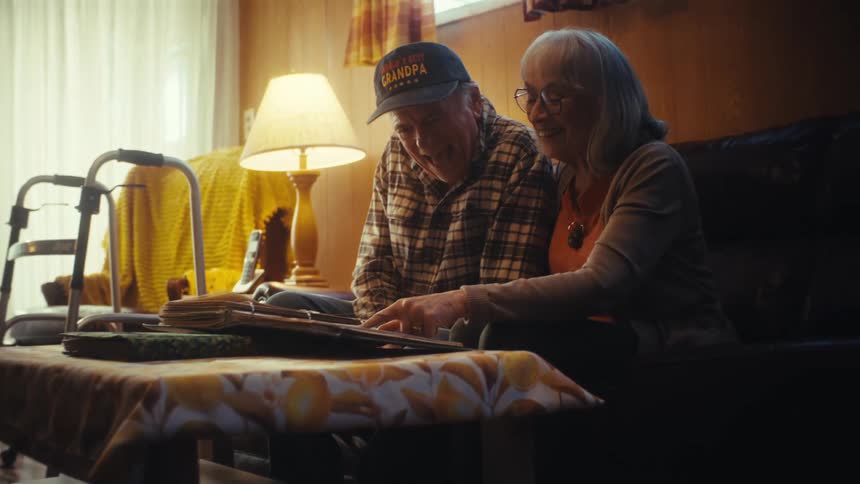 A loving elderly couple reminisces over a photo album, laughing as the grandfather excitedly dances in his seat