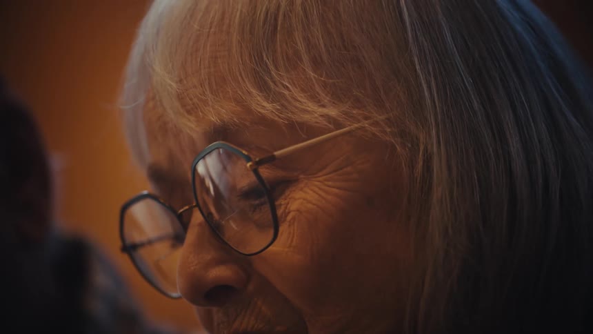 An extreme close-up profile shot of an elderly woman with glasses as she breaks into a gentle smile in a warmly lit room
