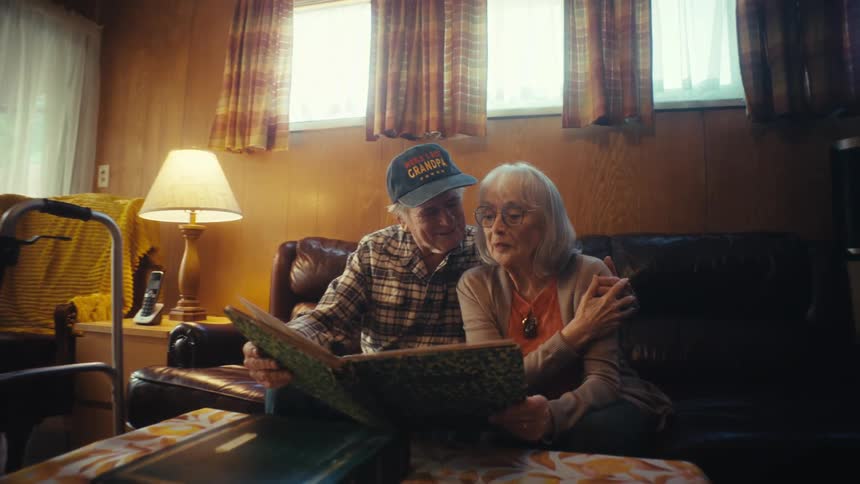 A heartwarming moment as an elderly husband and wife look through a photo album together, smiling and embracing on their living room couch